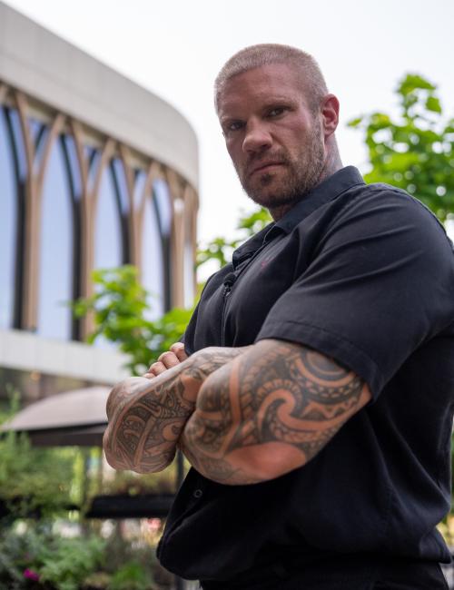 Getatoeëerde man in een zwarte blouse kijkt serieus in de camera met zijn armen over elkaar.